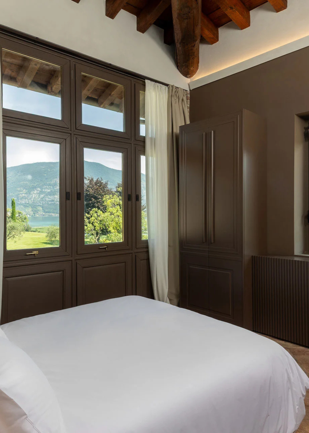 A modern bedroom interior featuring a large bed with white bedding, dark brown wooden wardrobes, and a multi-pane window offering a scenic view of the lake and mountains - one of the double rooms available at The Candle Cluub's Retreat Cluub