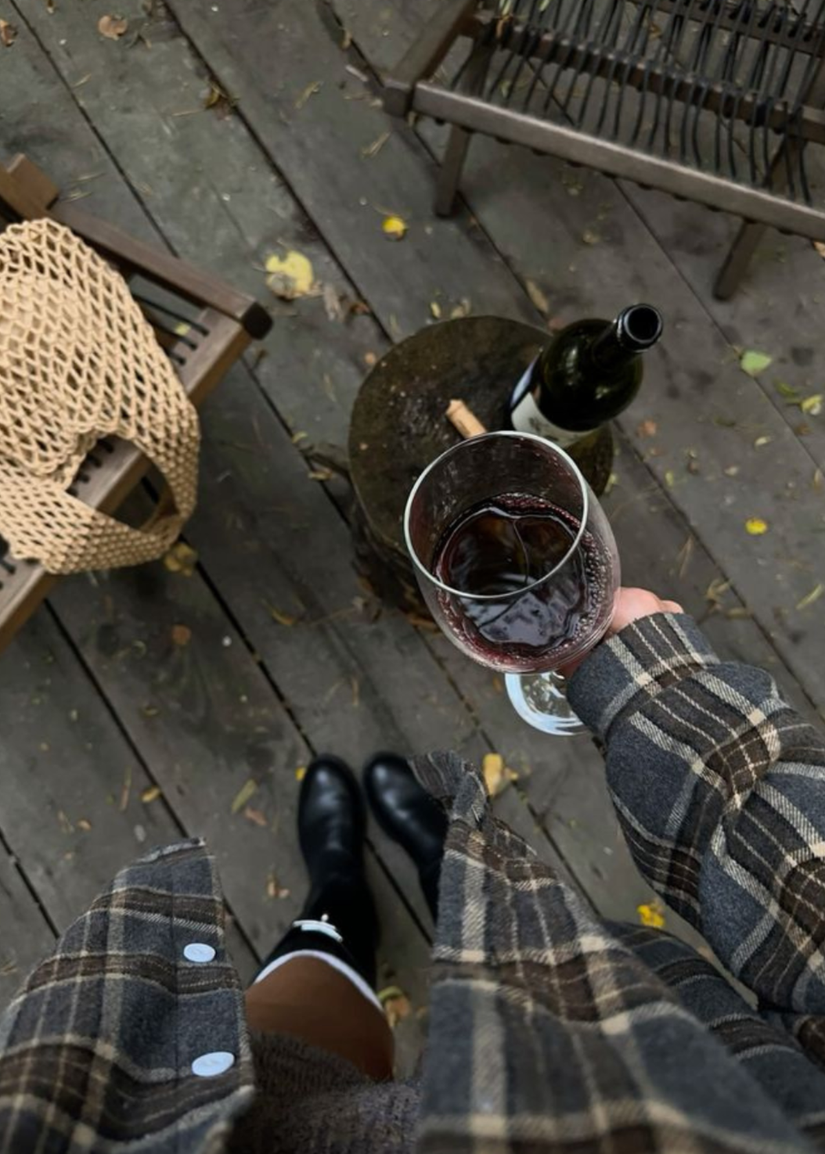 A top-down view of a person holding a glass of red wine on a wooden deck, wearing a brown and gray plaid flannel shirt and black rain boots ath The Candle Cluub Retreat Cluub