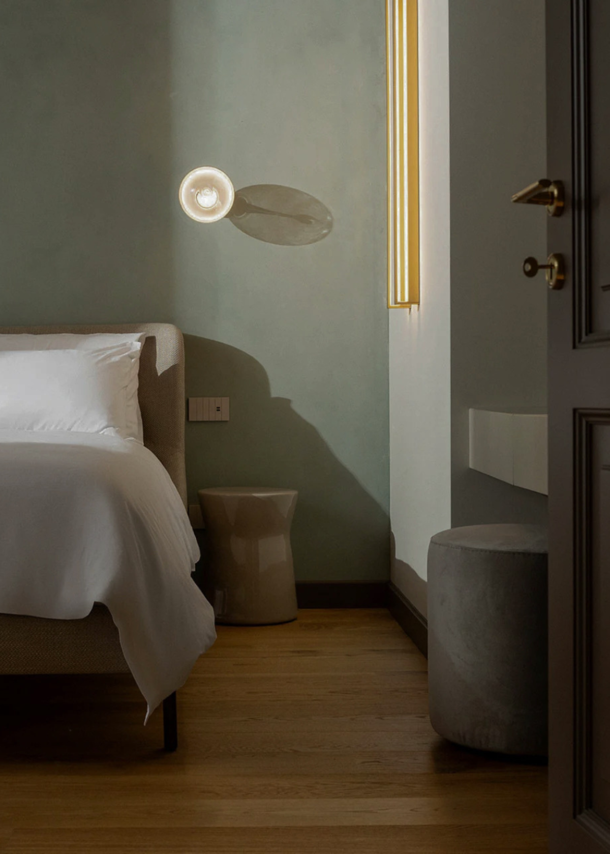 A modern bedroom interior with a bed dressed in white linens, a muted sage green wall, a sculptural wall light, wood flooring, and a dark upholstered stool as a room option for The Candle Cluub's Retreat Cluub