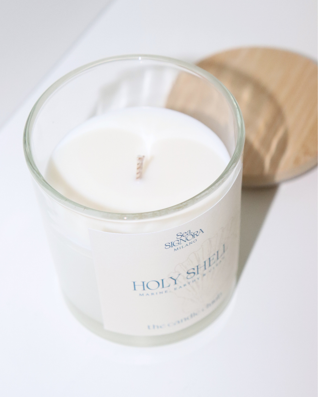 A white soy and coconut Candle Cluub candle in a clear glass jar with a white label reading 'Holy Shell' and 'The Candle Company,' photographed on a white surface with a light wood lid or coaster behind it.
