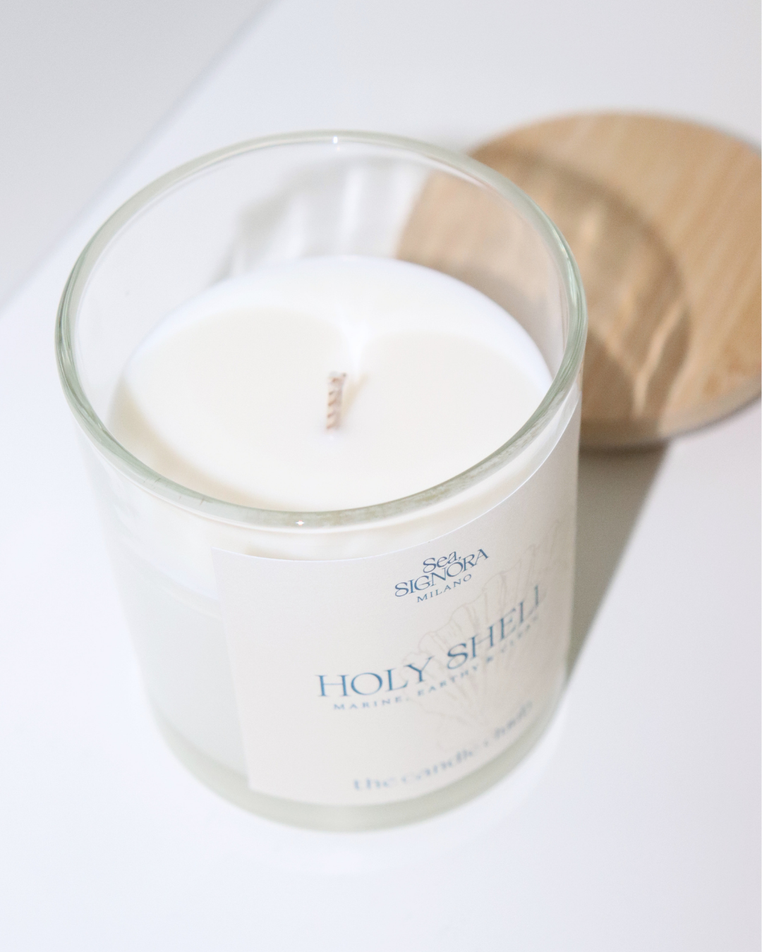 A white soy and coconut Candle Cluub candle in a clear glass jar with a white label reading 'Holy Shell' and 'The Candle Company,' photographed on a white surface with a light wood lid or coaster behind it.