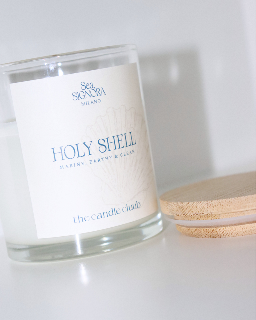 A Candle Cluub candle labeled 'Holy Shell' by Sea Sigorna Milano and The Candle Club, shown on a white surface with a bamboo lid beside it.