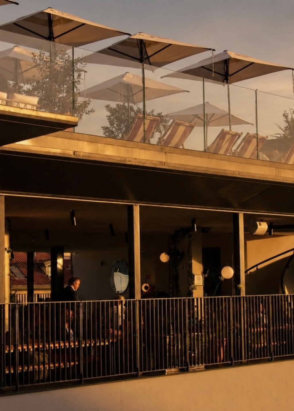 A modern outdoor terrace or rooftop lounge at golden hour, at Calimala Rooftop with large beige patio umbrellas, lounge chairs, glass railings, and warm sunlight casting long shadows across the structure for The Candle Cluub event