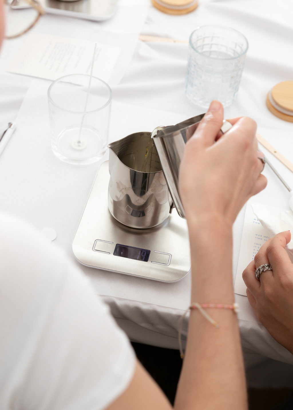 A close-up lifestyle shot of a person pouring candle wax from a stainless steel pitcher resting on a white digital scale over a countertop at The Candle Cluub workshop