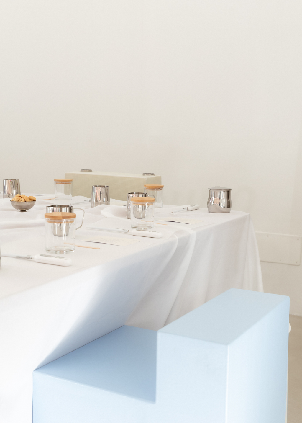 A styled dining table setup with a white tablecloth, clear The Candle Cluub jars with light wood lids, silver metal containers, and minimalist light blue and beige pedestal blocks in a bright, minimal room for The Candle Cluub workshop